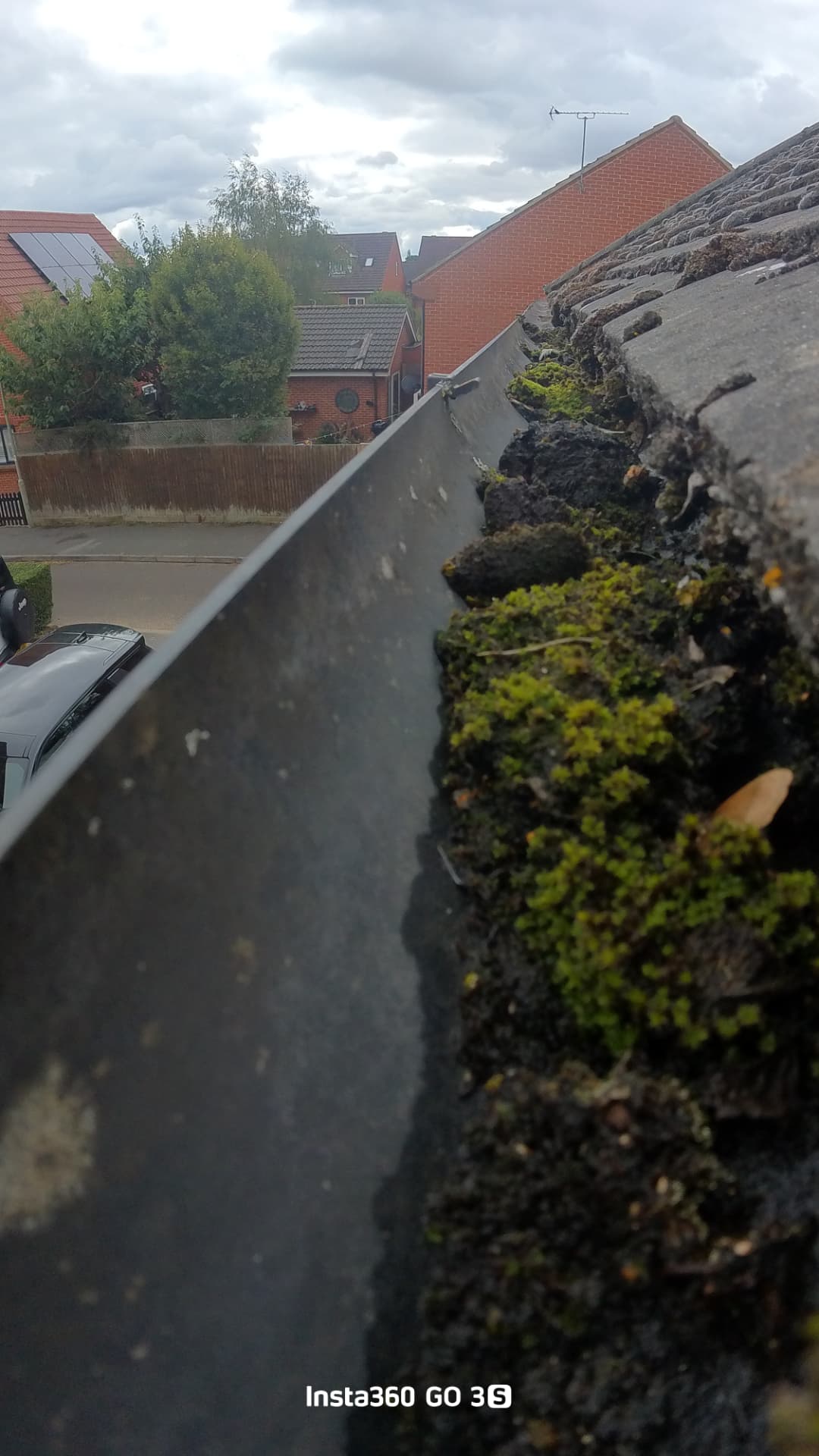 Insta360 camera view along a square-style gutter on Lea Park, Thame, with the gutter packed with moss beside the roof tiles and neighbouring brick houses in the background